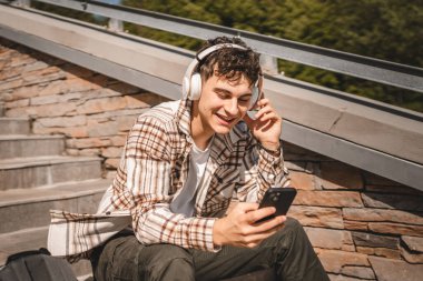 Genç mutlu adam merdivenlerde kulaklıkla oturur ve cep telefonuyla müzik dinler.