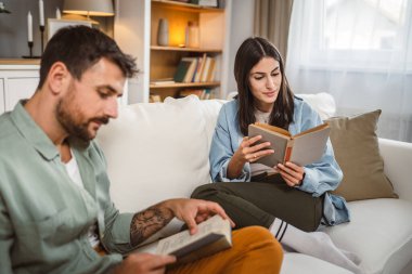Erkek ve kadın Kafkasyalı yetişkin çift evde kanepe yatağında kitap okuyor.