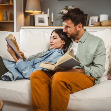 Erkek ve kadın Kafkasyalı yetişkin çift evde kanepe yatağında kitap okuyor.