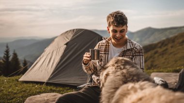 Delikanlı, çadırın önüne otur ve köpekle dağda kahve iç.