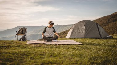 Kulaklıklı genç adam, meditasyon yap ve dağdaki çadırın önüne otur.