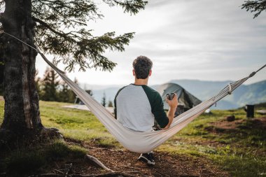 Genç çocuk bir hamakta oturur ve dağda kahve içer.