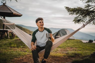 Genç çocuk dağda bir fincan kahveyle hamakta oturuyor.