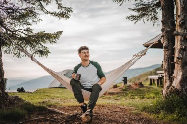 Genç çocuk dağda bir fincan kahveyle hamakta oturuyor.