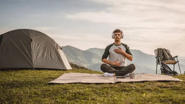 stock image Young boy with headphones, meditate and sit down in front of tent on the mountain