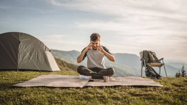 Genç çocuk dağdaki battaniyeye otur ve kulaklıkla ve cep telefonuyla müzik dinle.