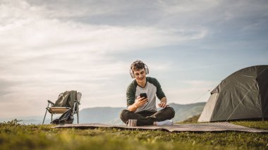 Genç çocuk dağdaki battaniyeye otur ve kulaklıkla ve cep telefonuyla müzik dinle.