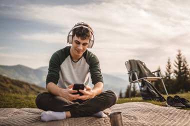 Genç çocuk dağdaki battaniyeye otur ve kulaklıkla ve cep telefonuyla müzik dinle.