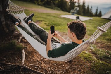 Genç çocuk hamakta uzanıyor ve dağda cep telefonu kullanıyor.