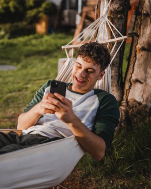 Genç çocuk hamakta uzanıyor ve dağda cep telefonu kullanıyor.