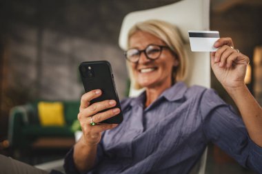 Evde çalışan olgun bir kadın cep telefonundaki kredi kartıyla internetten satın almak için ara verir.