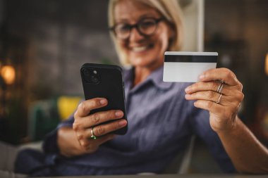 Evde çalışan olgun bir kadın cep telefonundaki kredi kartıyla internetten satın almak için ara verir.