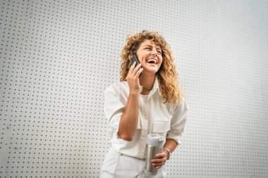 Yetişkin bir kadın cep telefonuyla konuşur termos bardağını sıcak içecekle tutar.