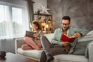 Sevgilisi evde tablet kullanırken erkek arkadaşı kitap okuyor.