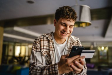 Genç çocuk ayakta duruyor ve kredi kartıyla cep telefonundan online alışveriş yapıyor.