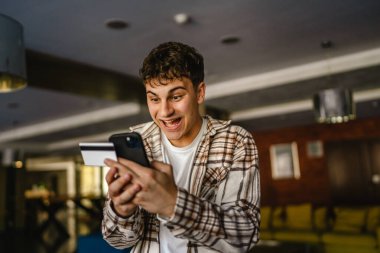 Genç çocuk ayakta duruyor ve kredi kartıyla cep telefonundan online alışveriş yapıyor.