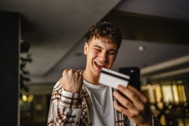 Genç çocuk ayakta duruyor ve kredi kartıyla cep telefonundan online alışveriş yapıyor.