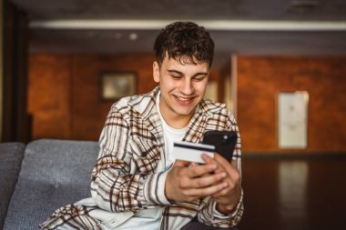 Genç çocuk ayakta duruyor ve kredi kartıyla cep telefonundan online alışveriş yapıyor.