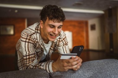 Genç çocuk ayakta duruyor ve kredi kartıyla cep telefonundan online alışveriş yapıyor.