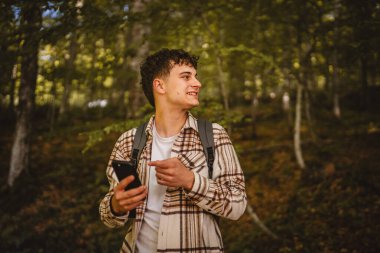 Sırt çantalı genç bir adam ormanda dolaşıyor ve yön tarifi ya da mesaj için cep telefonu kullanıyor, interneti keşfediyor.