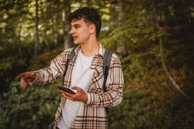 Sırt çantalı genç bir adam ormanda dolaşıyor ve yön tarifi ya da mesaj için cep telefonu kullanıyor, interneti keşfediyor.