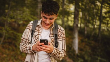 Sırt çantalı genç bir adam ormanda dolaşıyor ve yön tarifi ya da mesaj için cep telefonu kullanıyor, interneti keşfediyor.