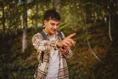 Sırt çantalı genç bir adam ormanda dolaşıyor ve yön tarifi ya da mesaj için cep telefonu kullanıyor, interneti keşfediyor.