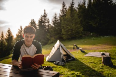 Genç yetişkin çocuk çadırın önünde oturur ve dağda bir kitap okur.