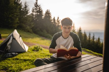 Genç yetişkin çocuk çadırın önünde oturur ve dağda bir kitap okur.