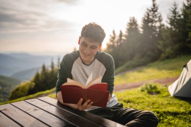 Genç yetişkin bir çocuk dağın tepesinde oturur ve dağda bir kitap okur.