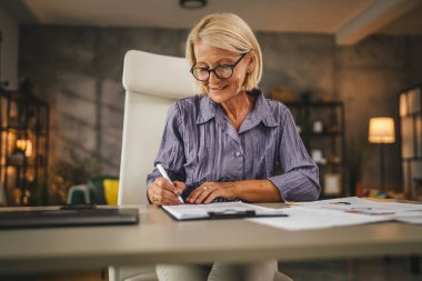 Olgun bir kadın evde çalışır ve sigorta imzalar, panoya bir belge koyar.
