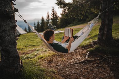 Genç bir çocuk dağdaki bir hamakta uzanıp kitap okuyor.