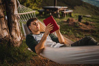 Genç bir çocuk dağdaki bir hamakta uzanıp kitap okuyor.