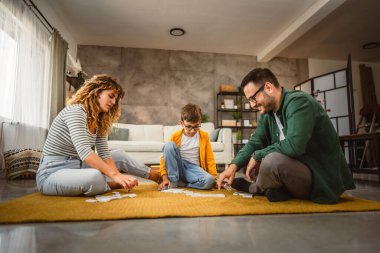 Baba, anne ve oğul oturma odasında yerde domino oynuyorlar.