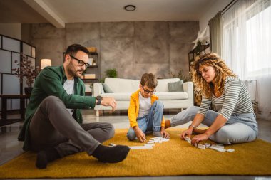 Baba, anne ve oğul oturma odasında yerde domino oynuyorlar.