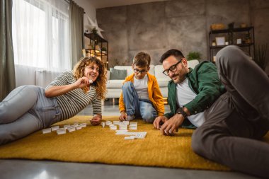 Baba, anne ve oğul oturma odasında yerde domino oynuyorlar.