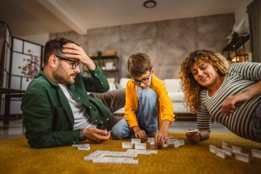 Baba, anne ve oğul oturma odasında yerde domino oynuyorlar.