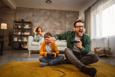 Anne ve babası joystickle video oyunu oynarken, anne karısı onları izliyor.