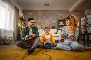 Anne onları desteklerken ve kahve içerken baba ve oğul joystick ile video oyunu oynuyorlar.