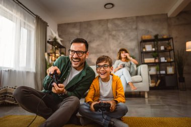Anne ve babası joystickle video oyunu oynarken, anne karısı onları izliyor.