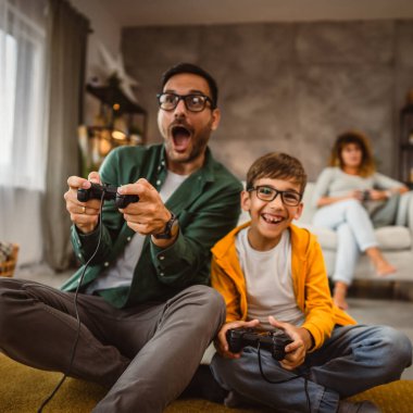 Anne ve babası joystickle video oyunu oynarken, anne karısı onları izliyor.