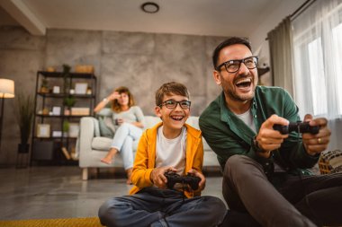 Anne ve babası joystickle video oyunu oynarken, anne karısı onları izliyor.