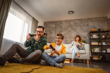 Anne ve babası joystickle video oyunu oynarken, anne karısı onları izliyor.