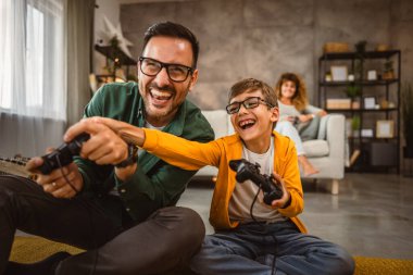 Anne ve babası joystickle video oyunu oynarken, anne karısı onları izliyor.
