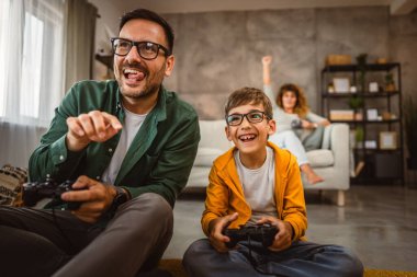 Anne ve babası joystickle video oyunu oynarken, anne karısı onları izliyor.