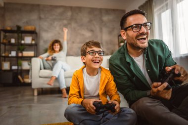 Anne ve babası joystickle video oyunu oynarken, anne karısı onları izliyor.