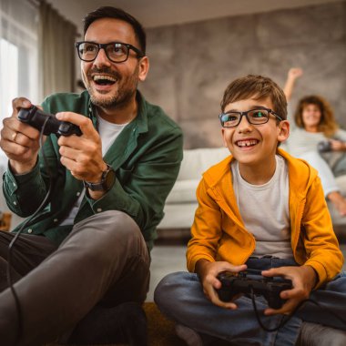 Anne ve babası joystickle video oyunu oynarken, anne karısı onları izliyor.
