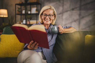 Olgun yaşlı kadın oturma odasında oturur kitap okur ve kahve içer.