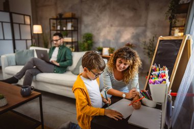 Anne ev ödevi yapar, oğul çizer, baba arka planda çalışır.