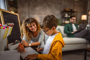 Anne ev ödevi yapar, oğul çizer, baba arka planda çalışır.